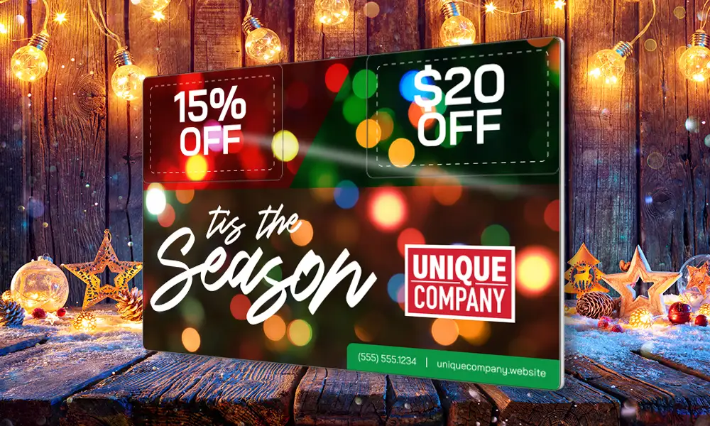 Holiday AcclaimMailer™ jumbo size direct mail plastic postcard, featuring two detachable offer cards, angled on a wooden counter with a blurred background.