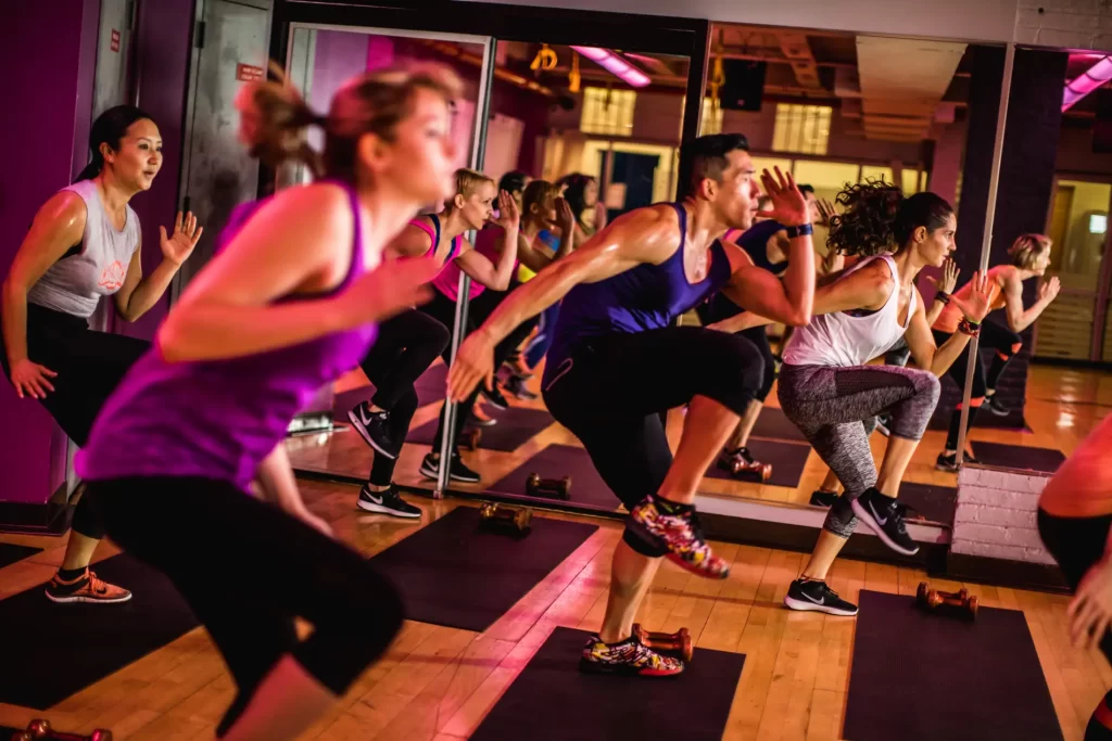Group fitness class performing high-intensity interval training (HIIT) exercises on black mats in a gym.