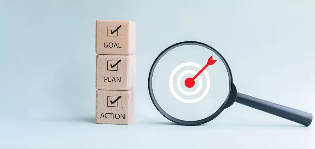 Goal, plan, and action concept showing wooden blocks with check marks and magnifying glass showing a target in the middle.