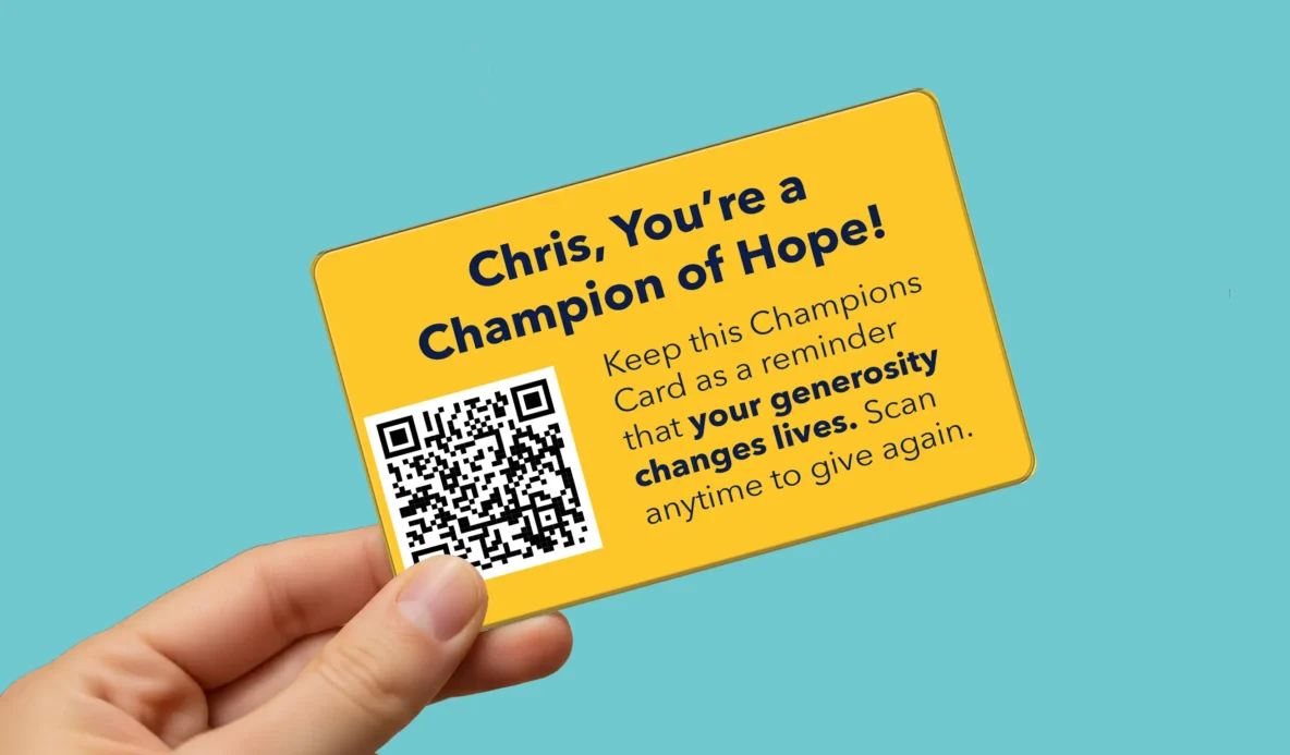 Hand holding the break off wallet card from a Champions of Hope nonprofit direct mail fundraising postcard featuring a QR code for easy donation.