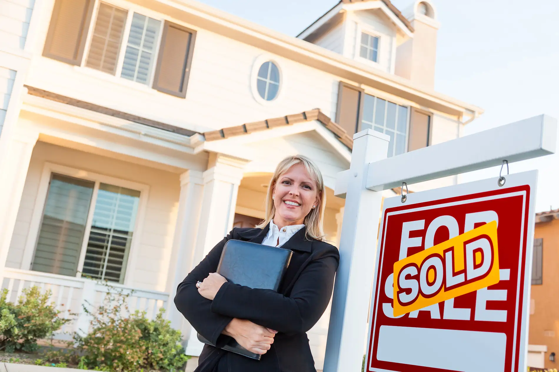 Female real estate agent standing in front of a house next to a "sold" sign